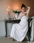 Tummahiuksinen nainen valkeassa yömekossa istumassa antiikkisen pöydän ääressä, levittämässä deovoidetta kainaloonsa. Taustalla lempeästi palavia mehiläisvahakynttilöitä ja valkoisia kukkia maljakossa.