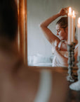 Peilikuva naisesta valkoisessa yömekossa, levittämässä deovoidetta kainaloonsa. Peilin edessä loistavat kauniit mehiläisvahakynttilät.