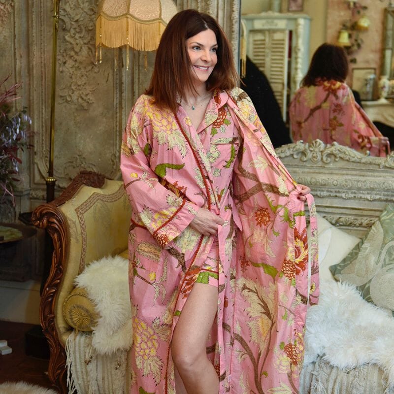 Hymyileväinen nainen seisoo Powell Craftin vaaleanpunainen kasviaiheinen aamutakki ja pyjamasetti päällään antiikkisohvan ja peilin edessä.