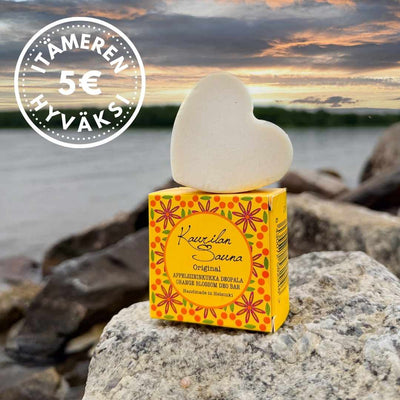 Kaurilan saunan appelsiininkukantuoksuinen deopala kauniin keltaisen pahvipakkauksensa päällä rantakivillä. Taustalla meri ja auringonlasku, vasemmassa yläkulmassa logo, jossa lukee "5€ Itämeren hyväksi", John Nurmisen säätiön yhteistyökampanjaan liittyen.