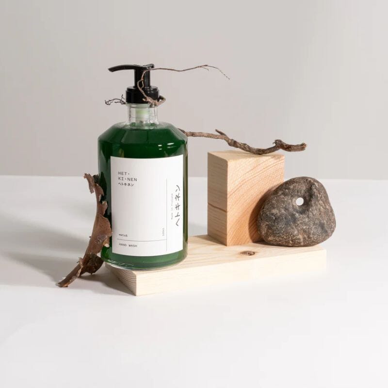 Hetkinen nestemäinen syvän vihreä käsisaippua 'Metsä' kirkkaassa pumppupullossa laudanpätkän päällä. Asetelmaan kuuluu myös kivi ja puunkappaleita.