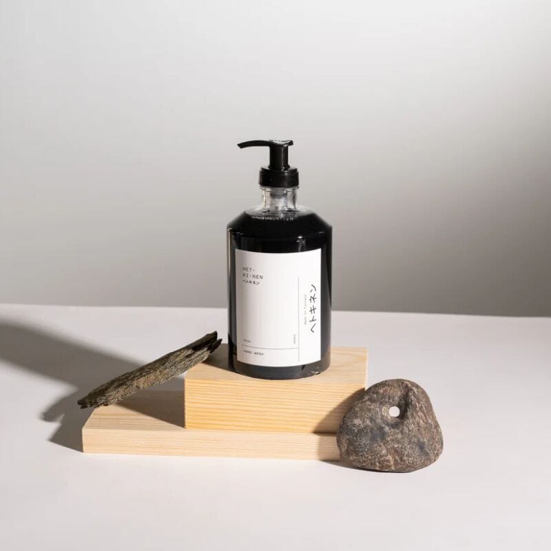 Hetkinen nestemäinen, mustaakin mustempi käsisaippua 'Sisu' kirkkaassa pumppupullossa, jossa tyylikäs valkoinen etiketti, ja joka on aseteltu laudanpätkien päälle kivenmurikoiden kanssa.