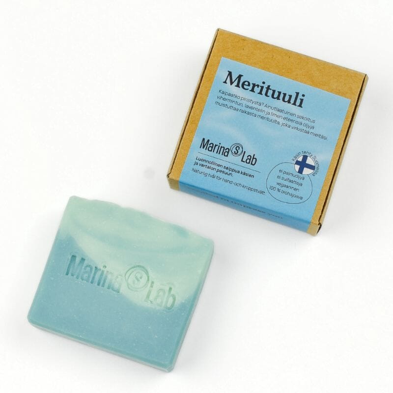 Marina's Lab Merituuli-palasaippua ja sen kartonkipakkaus valkoisella taustalla. Saippua on kaunis kaksisävyinen merensininen, pakkauksen etiketti taas muistuttaa taivasta.