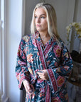 Powell Craftin puuvillainen kimono-aamutakki, jossa männynvihreällä pohjalla punaisen-, pinkin- ja sinapinsäyisiä block print -kuvioita ikkunasta ulos katsovan naisen yllä.