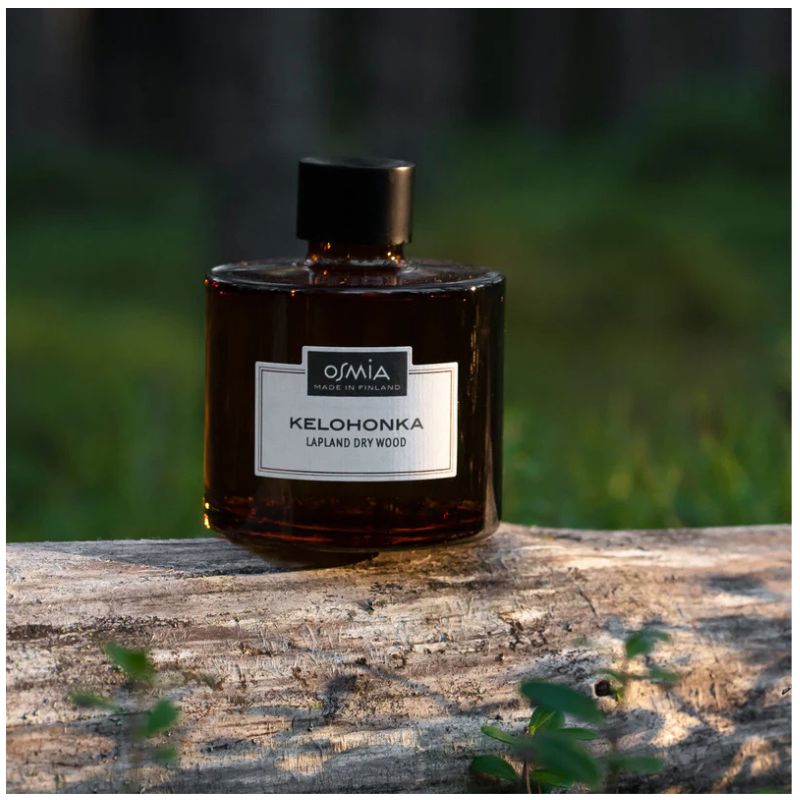 Osmian Kelohonka-huonetuoksu tyylikkässä ruskealasisessa 250ml pullossa, paistattelemassa kelohongan päällä ilta-auringossa.