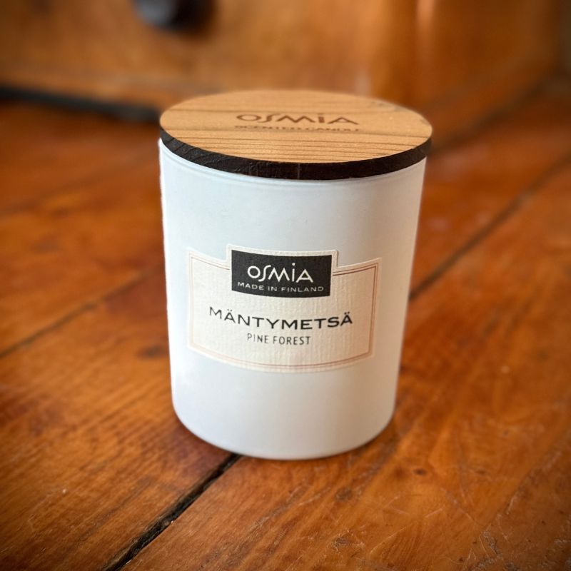 Osmia Mäntymetsä -tuoksukynttilä valkoisessa lasipurkissa puu kannella, kuvattuna lämpimässä valaistuksessa vanhalla puisella lattialla. 