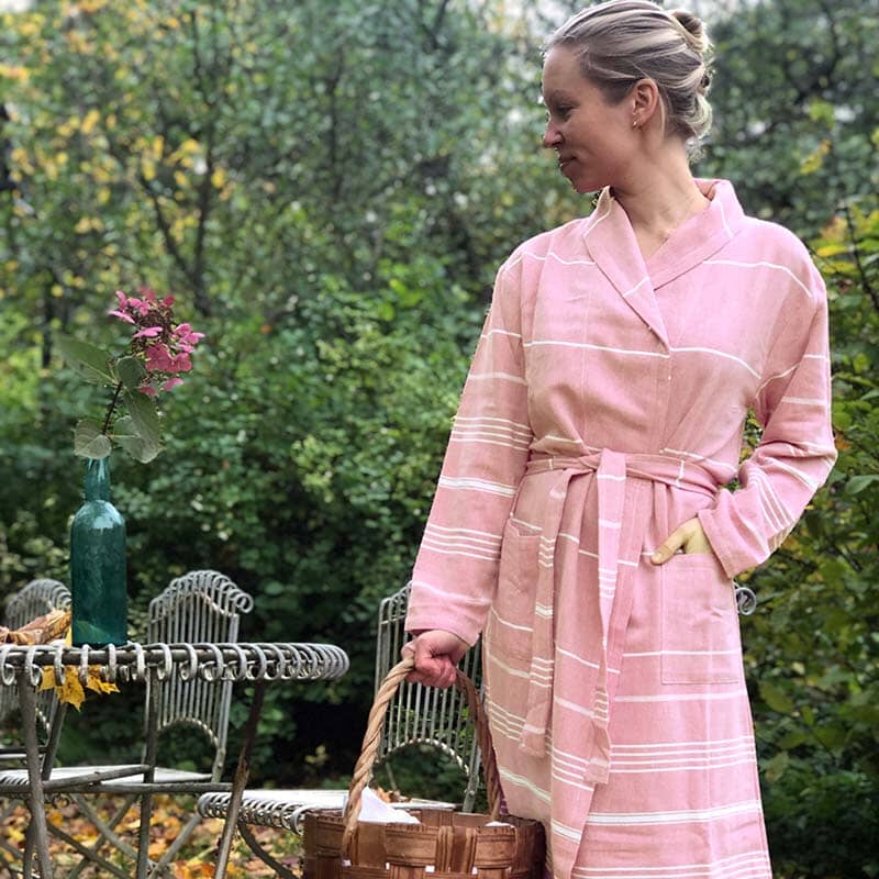 Saunatontun vaaleanpunainen valkoraitainen Roosan hamam-kylpytakki syksyisessä puutarhassa seisovan naisen yllä.