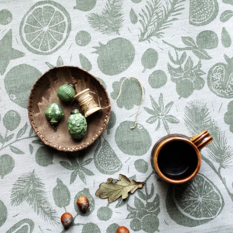 Lapuan Kankurit Talven Tuoksut -kangas, jonka päällä Arabian vintage-muki, tammenterhoja ja joulukoristeita pienellä ruskealla lautasella.