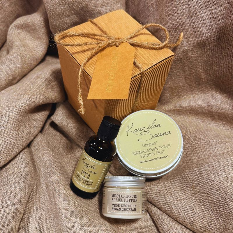 Kaurilan Saunan Tontun Saunasetti 2 - Lahjapaketti vaaleanruskean kankaan päälle aseteltuna. Lahjapaketti on tyylikästä luonnollisen ruskean sävyistä aaltokartonkia ja se on sidottu juuttinarulla, johon on kiinnitetty samaa sävyä oleva pakettikortti, jossa lukee kultaisella 'Hyvää joulua'. Paketin vieressä on siihen kuuluvat Turve-löyöytuoksu, Suomalainen turvenaamio ja Vegaaninen mustapippuri-deovoide.