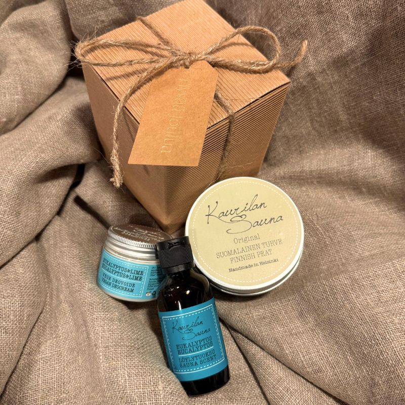 Kaurilan Saunan Tontun Saunasetti Ⅲ - Lahjapaketti vaaleanruskean kankaan päälle aseteltuna. Lahjapaketti on tyylikästä luonnollisen ruskean sävyistä aaltokartonkia ja se on sidottu juuttinarulla, johon on kiinnitetty samaa sävyä oleva pakettikortti, jossa lukee kultaisella 'Hyvää joulua'. Paketin vieressä on siihen kuuluvat Eukalyptus-löyöytuoksu, Suomalainen turvenaamio ja Vegaaninen eukalyptus & lime -deovoide.