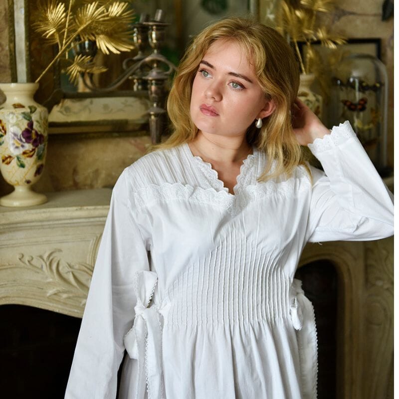 Nuori nainen seisoo vanhan takan edessä mietteliäänä, yllään valkoinen Virginia-yöpaita yritykseltä Powell Craft.