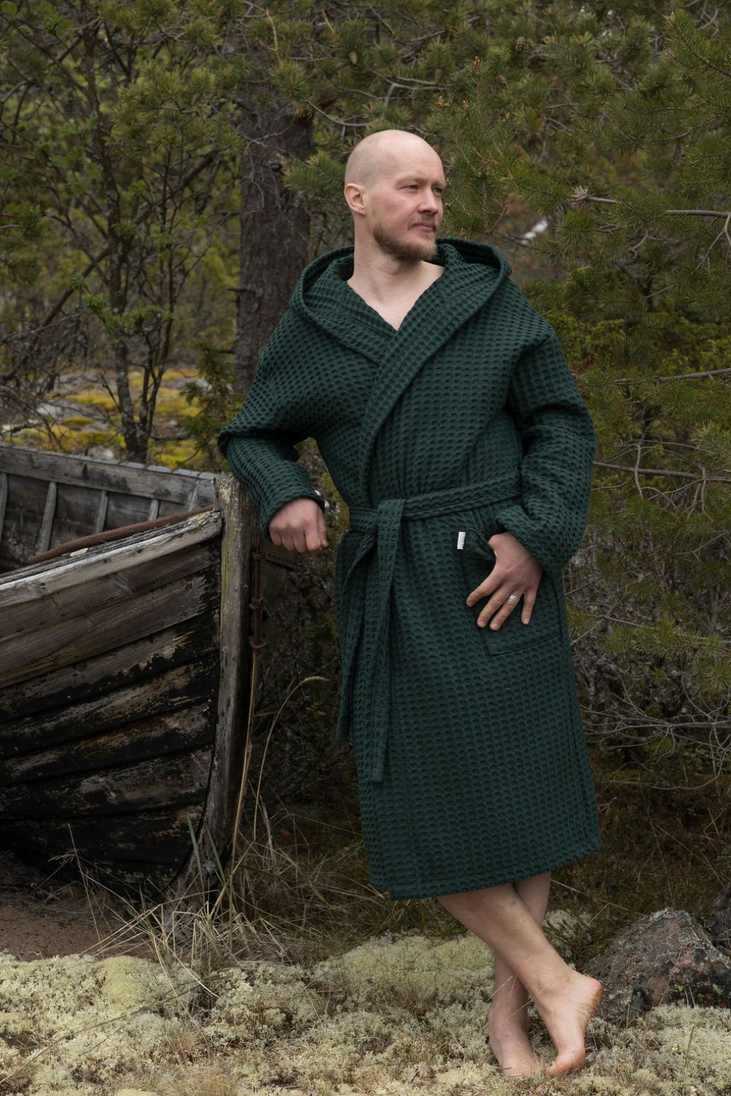 Metsänvihreään Keltainen designin vohvelikylpytakkiin pukeutunut mies nojailee metsässä vanhaan puuveneeseen.