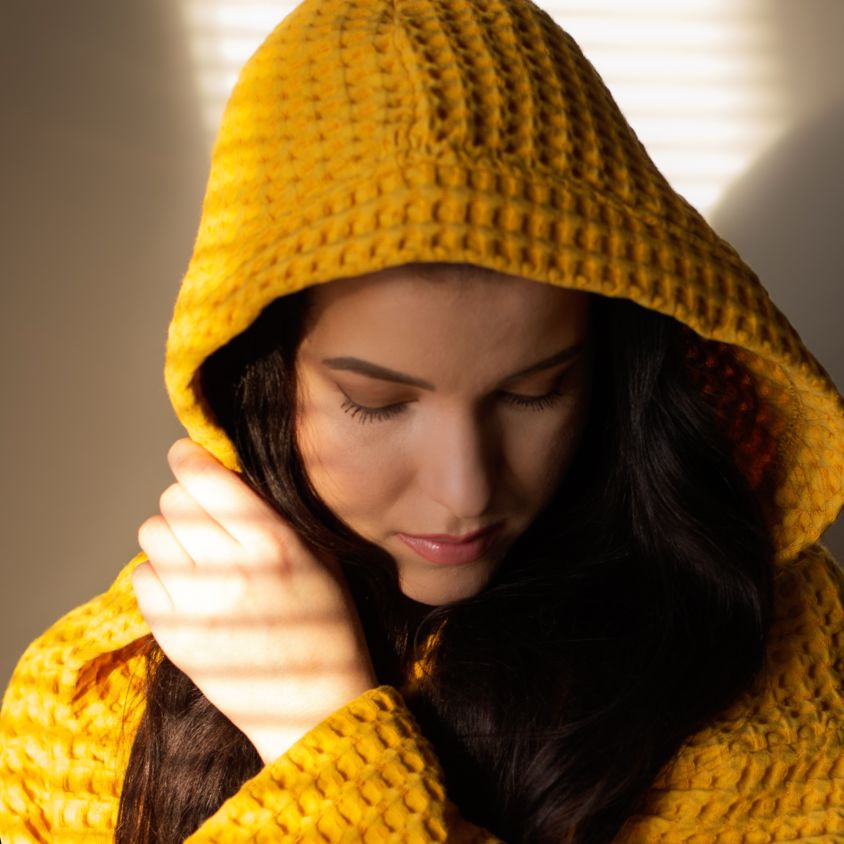 Okraan Keltainen designin vohvelikylpytakkiin sonnustautunut nainen mietteliäänä sälekaihtimien läpi paistavan auringon valossa.