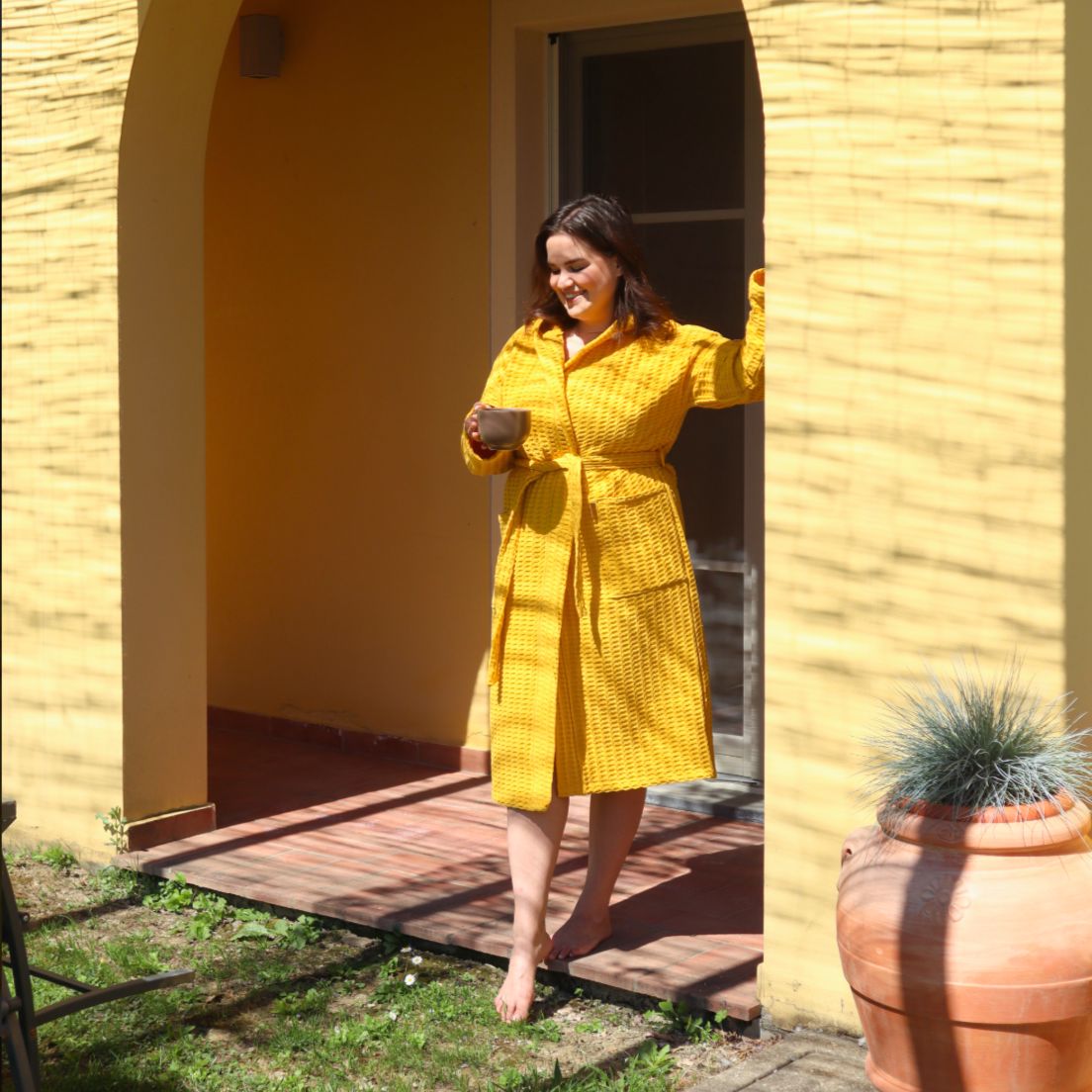 Okrankeltaiseen Keltainen designin vohvelikylpytakkiin pukeutunut nainen astuu ovensuusta paljain varpain nurmikolle, kahvikuppi kädessään.