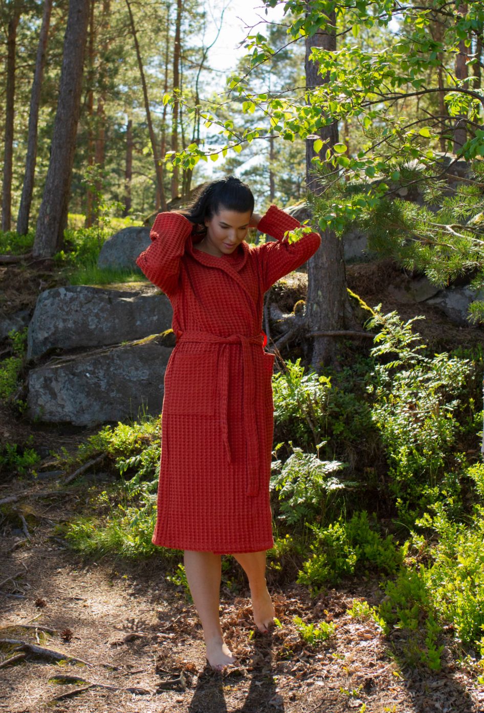 Tummahiuksinen nainen käyskentelee kesäisessä metsässä, yllään Keltainen designin tiilenpunaunen vohvelikylpytakki.