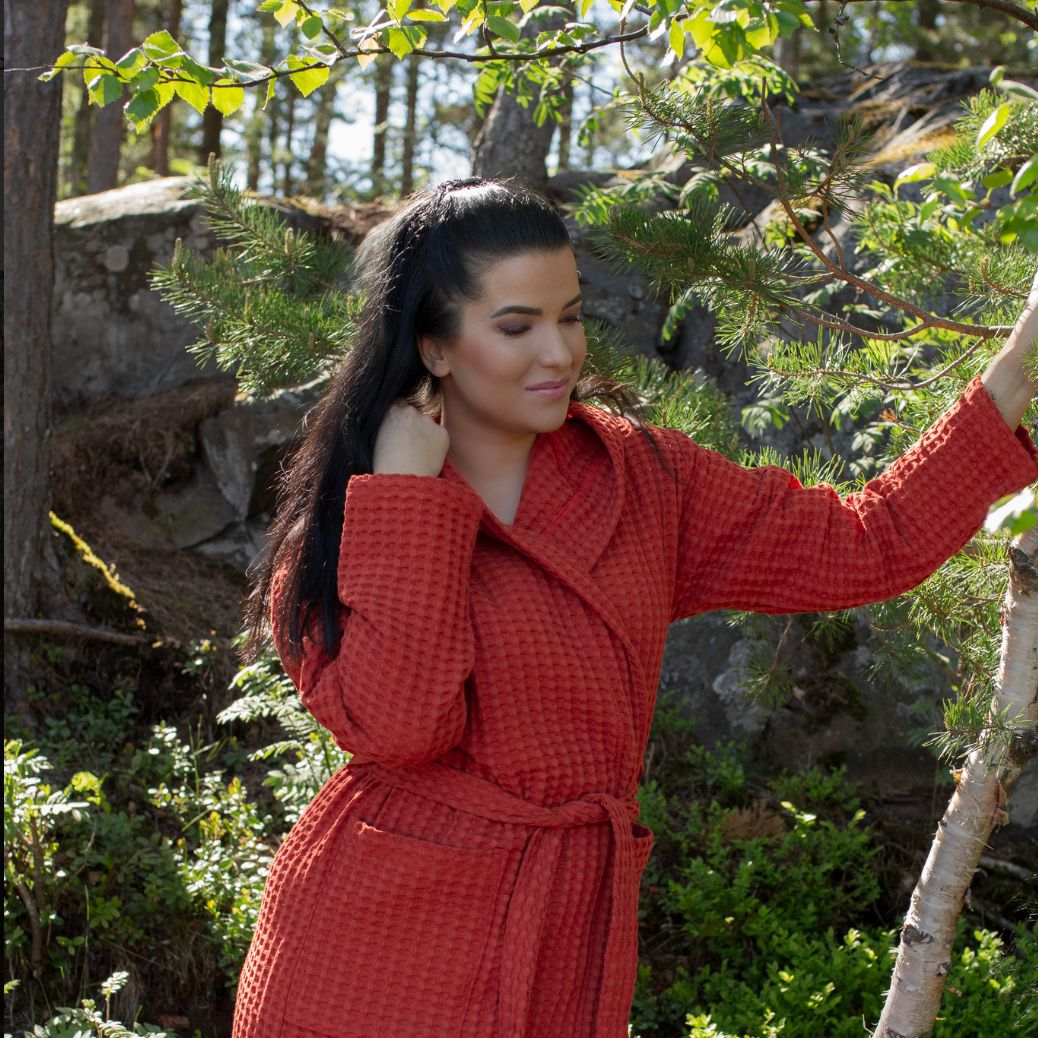 Tummahiuksinen nainen nojailee metsässä puuhun, yllään Keltainen designin tiilenpunaunen vohvelikylpytakki.