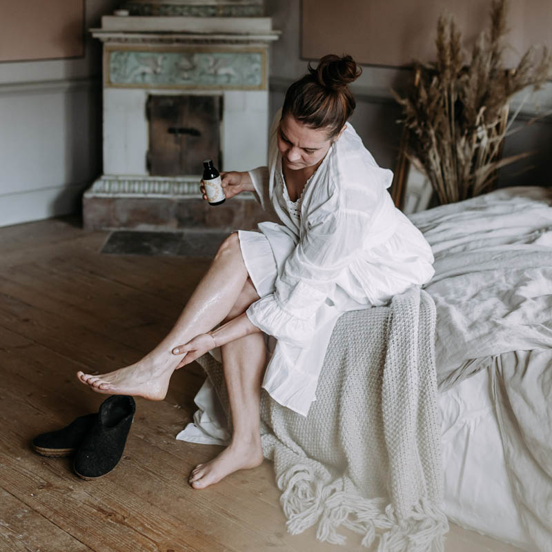 Tummahiuksinen, valkoisiin puuvillavaatteisiin pukeutunut nainen istumassa sängyn laidalla, hieromassa Kaurilan saunan appelsiinintuoksuista vartaloöljyä säärilleen. Taustalla vanha kakluuni ja maljakollinen kaisloja.