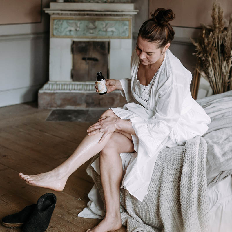 Sängyn reunalla istuva nainen levittämässä Kaurilan saunan vartaloöljyä säärilleen. Valkoiseen pitsiyöpaitaan ja aamutakkiin sonnustautuneen naisen edessä on mustat tohvelit, taustalla koristeellinen vanha kakluuni.