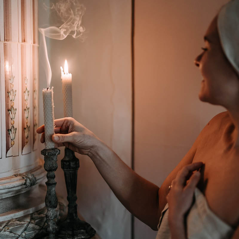 Pellavapyyhkeisiin pukeutunut nainen sammuttamassa Kaurilan saunan vaaleita mehiläisvahakynttilöitä. Sammuneesta kynttilästä nousee savukiehkuroita.