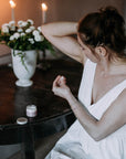 Valkoiseen yömekkoon pukeutunut nainen levittämässä Kaurilan Saunan vegaanista deovoidetta kainaloihinsa tumman antiikkisen pöydän ääressä. Taustalla käsintehtyjä mehiläisvahakynttilöitä ja valkoisia kukkia maljakossa. 